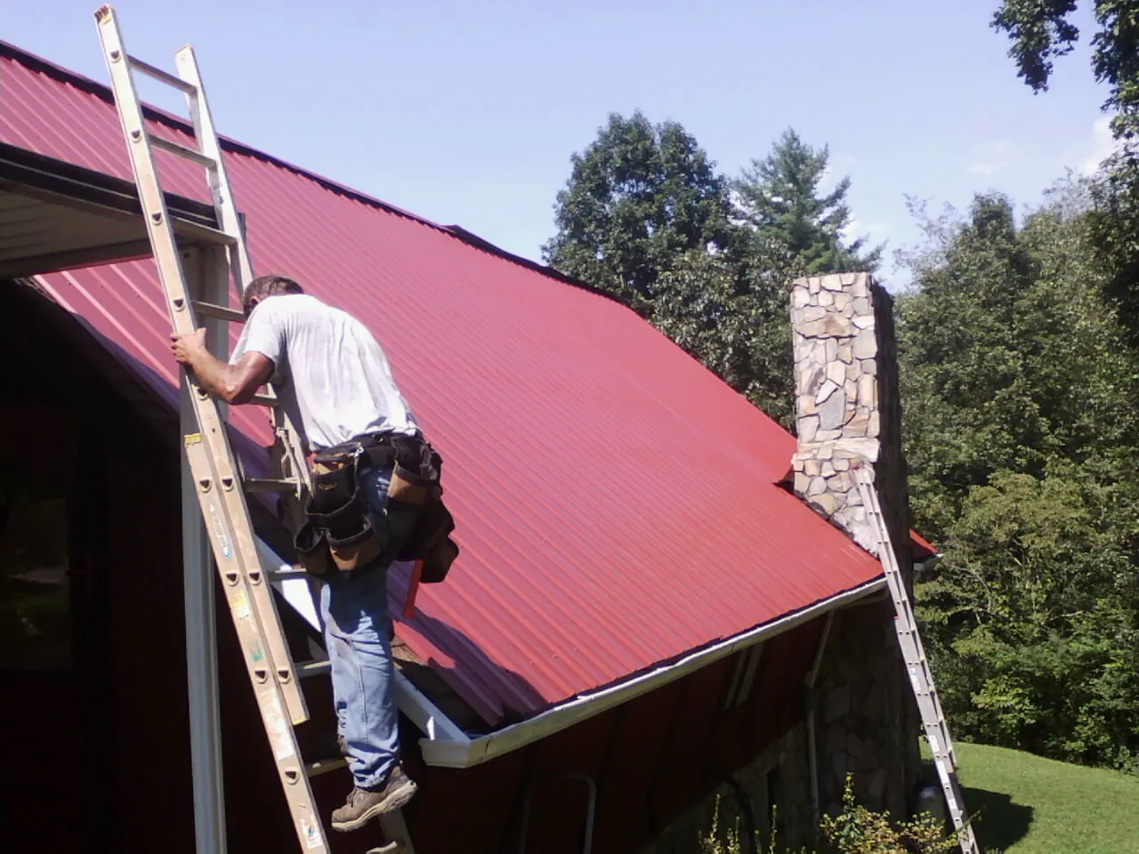 Roofer on a ladder beside a red metal roof and stone chimney — Soffit & Fascia Repair by Roofing Brothers in Tuscaloosa